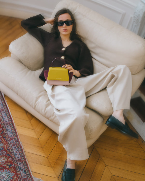 Sac ALEXIA NANO Colourblock Vesper tenu par la poignée, porté à la main, assise sur un canapé