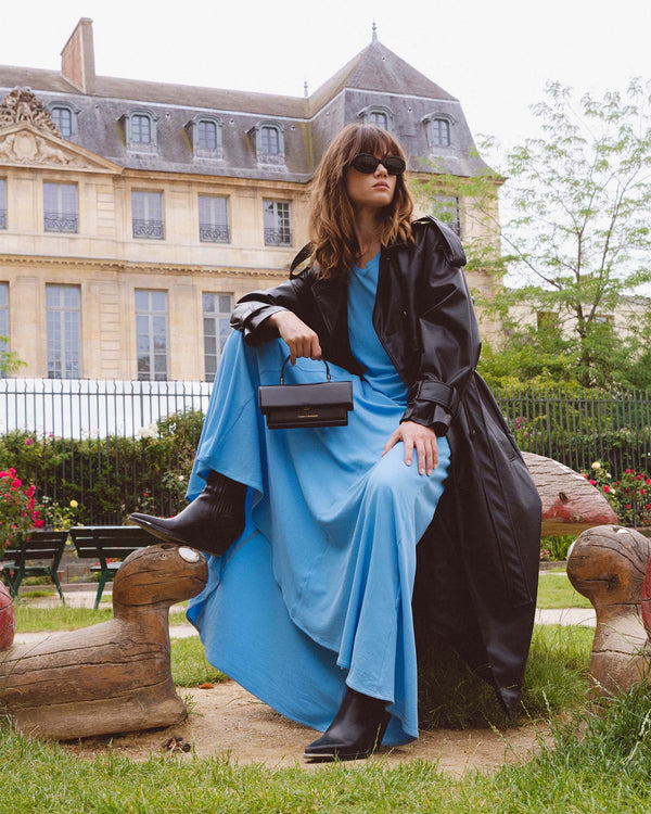 Sac THEA MINI noir tenu à la main par une femme assise en robe bleue et trench noir