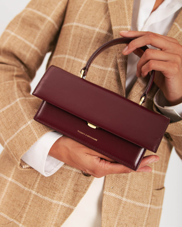 Sac THEA MINI Mahogany tenu à la main par son anse, fermoir doré, porté devant un blazer à carreaux