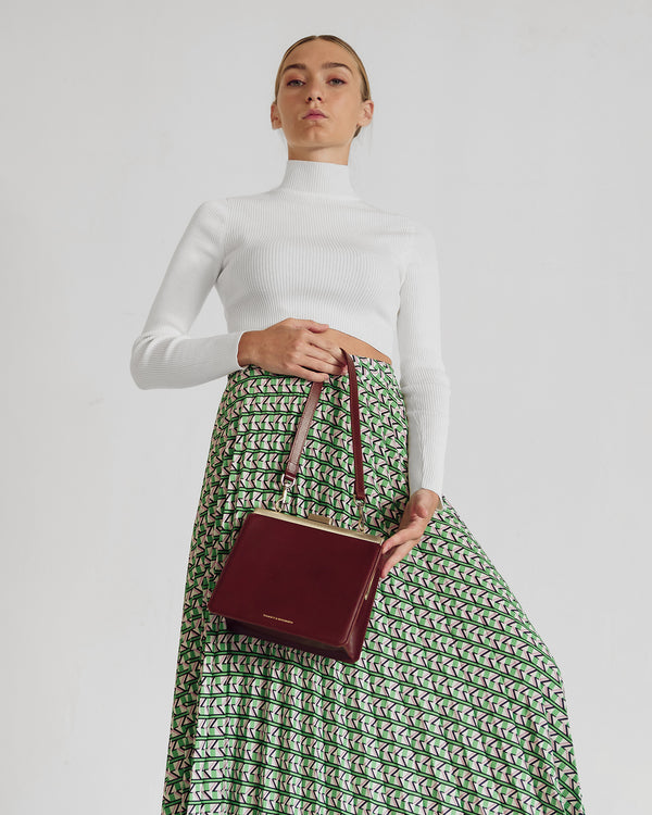 JEANNE bag in brown held by the handle, worn in front of a green printed skirt